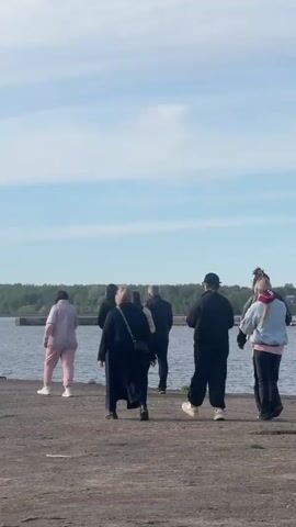 A group of approximately eight people is walking away from the camera on a concrete pier. They are walking towards a body of water with a distant treeline on the opposite shore. The sky is light blue with some wispy clouds. Most of the people are wearing dark clothing, though one is in a light pink tracksuit and another has a light denim jacket. The people are walking at a leisurely pace.