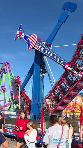 This video displays an amusement park ride with riders in suspended cages that swing back and forth. The ride is tall and blue with American flag decals. Several people are standing around the ride, some watching. Other rides and stalls are visible in the background. The sky is clear and blue.