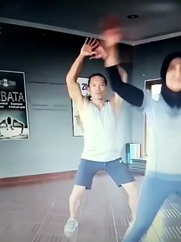The video shows two people exercising in a room with a dark grey wall. There are posters on the wall, one of which says "ZUMBA". The person on the left is a man wearing a white tank top and blue shorts. He is in the middle of a jumping jack motion with his arms extended to the sides. The person on the right is a woman wearing a light blue hijab and a light blue athletic shirt with long sleeves and matching blue leggings. She is also exercising, appearing to be performing similar movements to the man. The floor is tiled with a light brown and white pattern. The lighting is somewhat dim, and the image quality is grainy. The overall impression is of a fitness class or workout session.