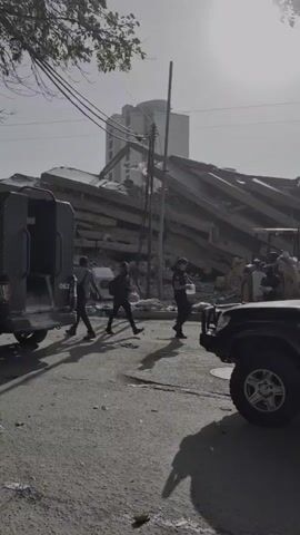 The video shows a street scene in what appears to be an urban area that has experienced significant destruction. In the background, a tall building stands, partially obscured by a massive pile of rubble, which looks like the collapsed remains of a multi-story structure. The debris consists of concrete slabs, beams, and other construction materials.

In the foreground, several vehicles are present. On the left, a dark-colored vehicle, possibly an armored van or truck, is parked facing away from the camera. Its side has markings, including "062". To the right and partially in frame, another dark-colored SUV or jeep is parked, with its front wheel and bumper visible. Between these vehicles, a man wearing a dark jacket and backpack walks across the street. Further into the scene, near the rubble, two people are visible, one wearing a dark jacket and backpack, and another person behind them. A yellow construction vehicle, possibly a backhoe or loader, is also present amidst the debris.

Scattered on the ground are various discarded items, including what appears to be a white washing machine or dryer, plastic sheeting, and other debris. Electrical wires hang precariously from poles in the middle ground, with some appearing damaged. The overall atmosphere is one of devastation and ongoing recovery or assessment efforts. The lighting suggests it is daytime, with a bright sky.