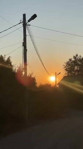 A child walks down a dirt road during sunset. The sun is low in the sky, casting a warm glow and lens flare across the scene. The child is silhouetted against the sunset, wearing a green t-shirt and a baseball cap. Overhead, telephone wires are strung between poles, with one pole prominently featured in the foreground. Trees and bushes line the road, and a distant house is visible behind the trees. The overall atmosphere is peaceful and serene.