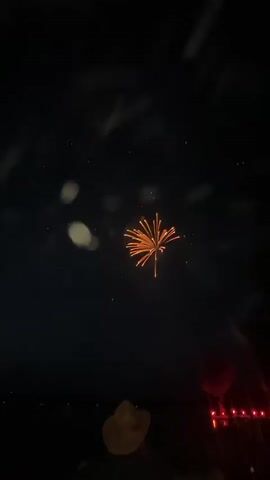 The video captures a night sky illuminated by fireworks. The fireworks are orange and yellow in color, exploding in a radiant pattern. In the foreground, silhouetted figures of people are visible, one wearing a cowboy hat. Red string lights are also present in the lower right corner of the frame, possibly part of a decoration or event setup. The overall environment appears to be an outdoor gathering for a fireworks display.