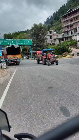 A red tractor with a blue canopy is pulling a trailer loaded with green foliage down a paved road. The tractor is on the right side of the road, moving towards the camera. In the background, there are several multi-story buildings situated on a hillside covered with trees. A green and white road sign is visible on the left side of the road. A yellow and red truck is parked on the left side of the road, facing away from the camera. The sky is overcast. The video is captured from the perspective of someone riding a motorcycle.