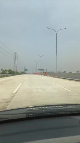 The video shows a view from inside a car driving on a highway under a cloudy sky. The highway is paved with concrete and has multiple lanes. Traffic cones and construction signs are visible, indicating road work in progress. Several cars are driving ahead. On the left side of the highway, there are tall power transmission towers and some greenery. The car's windshield wipers are visible at the bottom of the frame.