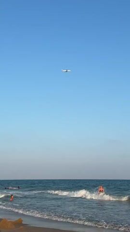 In this media file, a clear blue sky dominates the upper portion, with a distant airplane soaring high above. Below, the scene transitions to a tranquil beach setting at what appears to be late afternoon or early evening, given the soft lighting. Gentle waves roll onto the sandy shore. Several individuals are enjoying the water, with some wading in the shallows and others further out. One person in the foreground, standing near the water's edge, is wearing a black top and patterned shorts, while another person in the background is visible in the water. The overall atmosphere is one of leisure and relaxation, with people unwinding at the seaside. The sandy beach in the foreground features a small, sandcastle-like structure. The location is identified as Gavà, Spain. There is no visible text in the media file.