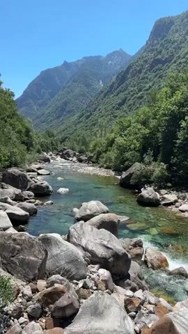 The video shows a scenic mountain valley with a clear river flowing through it. The river is surrounded by large rocks and lush green trees on the steep valley slopes. The sky is clear blue, indicating a sunny day. The camera is positioned at a low angle, looking downstream. The water in the river is turquoise and appears to be moving swiftly in some areas, with small rapids visible. The mountains in the background are covered in dense vegetation, with some rocky outcrops and peaks visible. The overall impression is one of natural beauty and tranquility.
