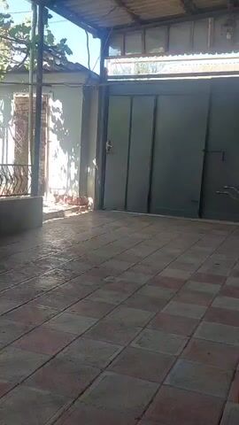 The video shows an outdoor area with tiled flooring. To the left, there is a white wall with a brown trim, and a metal railing. A tiled structure with a faucet and a sink is visible, as well as a broom leaning against it. Further to the left, a pair of flip-flops is hanging from a railing. In the background, there are trees and vegetation. To the right, a large metal gate is visible. The lighting suggests it is daytime.