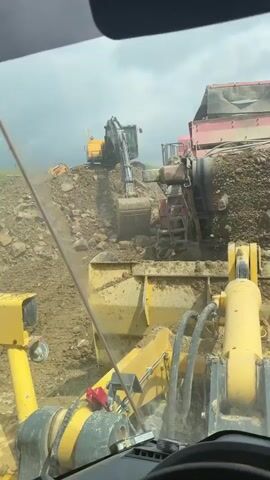 The video shows a construction site on a cloudy day from the perspective of someone inside the cabin of a yellow heavy machinery vehicle. The vehicle is facing a large, reddish-pink soil screener that's processing dirt and rock. An excavator is on a pile of rubble next to the screener, loading it with more material. Another piece of yellow heavy machinery is in the background on the left. The windshield wipers in the heavy machinery vehicle are visible, though they are not in motion.