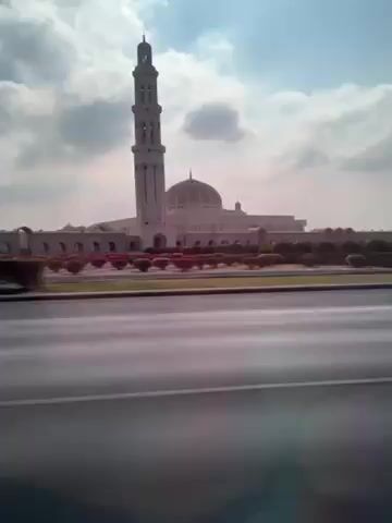 The video shows a view from a moving vehicle, likely a car, traveling on a paved road. The focus of the shot is a large, ornate mosque visible on the right side of the road. The mosque features a tall, slender minaret and a central dome, with a series of arches connecting the various parts of the building. A row of flowering bushes lines the median between the road and the mosque. The sky is partly cloudy with bright sunlight. As the car moves, the background shifts to include trees and distant mountains.