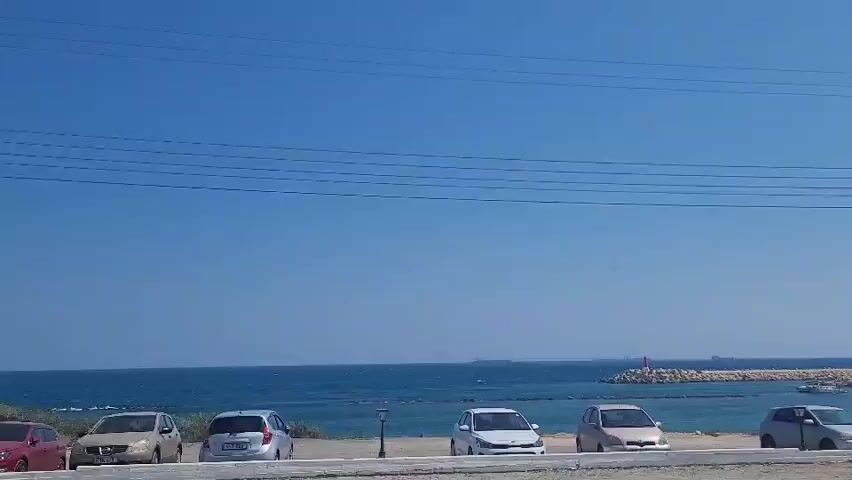 The video shows a blue sky with thin power lines running horizontally across it. Below the sky, the deep blue sea stretches out, meeting a rocky shore. On the shore, several cars are parked in a line. The cars are of various colors, including white, silver, light beige, and a darker color. Some of the cars are small hatchbacks. A few streetlights are visible between the cars. Further out, there's a breakwater made of large rocks, with a red-capped lighthouse at its end. Beyond the breakwater, several ships are visible on the horizon. The overall scene is that of a sunny day at a coastal parking area.