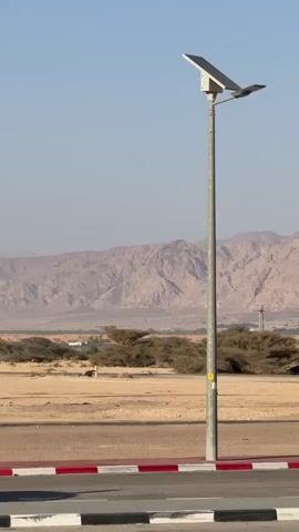The video shows a desert landscape with a mountain range in the background under a clear, pale blue sky. In the foreground, a road with a painted curb separates the asphalt from a sandy area with sparse vegetation. Several tall street lights are visible along the roadside, one of which is solar powered.
