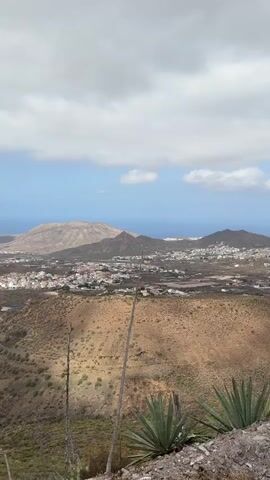 The video shows a panoramic view of a valley with a town or city spread across it. In the foreground, there are some dry plants and rocky ground. The middle ground consists of a dry, hilly landscape with patches of vegetation and some signs of human activity, possibly agriculture. In the background, the town is visible with numerous buildings, and beyond that, the blue ocean stretches to the horizon. Mountains are scattered across the landscape. The sky is partly cloudy with a mix of blue sky and white clouds. The lighting suggests it is daytime, likely overcast or with intermittent sunlight. There are no people or animals visible in the video. The camera appears to be mounted on a tripod or steady surface, and the view is static. The scene depicts a rural or semi-rural landscape transitioning into a coastal urban area.