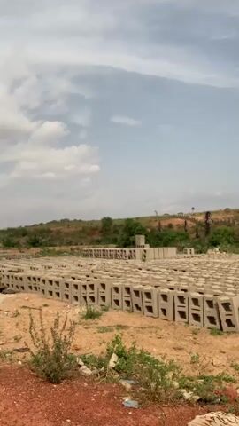 The video shows a field covered in concrete blocks under a partially cloudy sky. The blocks are arranged in rows, each containing several blocks with hollow rectangular openings. The ground is mostly bare dirt, with some sparse vegetation scattered throughout. In the background, there is a slightly elevated area with trees and some structures, possibly buildings, visible in the distance. There are no people or animals visible in the video.