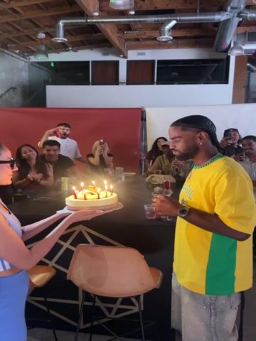 An indoor birthday celebration is captured in the video, with a man in a yellow and green shirt, featuring "BRASIL" text, standing to the right. He has braided hair and is seen with his hands clapped together as if making a wish, looking down at a birthday cake. A woman, partially visible in a blue top, holds the cake with several lit candles on a dark bar counter. In the background, a group of people are gathered, clapping, smiling, and recording the event with their phones, indicating a joyful atmosphere. The setting is a modern event space, possibly a studio or loft, with exposed industrial-style ceiling elements and lighting. The overall scene is bright, suggesting it is daytime.