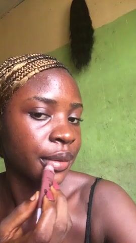 A person with brown skin and blonde-tipped braids is applying a light red lip product. They are wearing a black tank top. The wall behind them is green and a dark curly wig is hanging on the wall. They are looking at the camera. They purse their lips and look at the camera again.