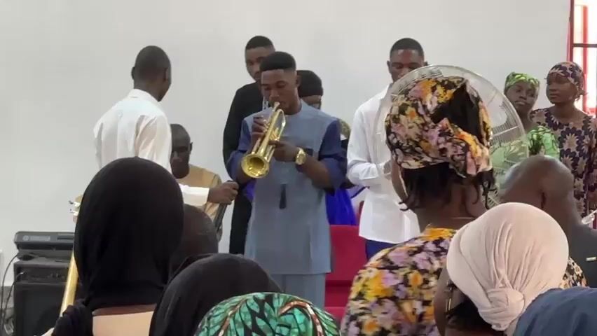 A young man in a light blue and dark blue two-tone long-sleeved tunic plays a brass trumpet in a room with white walls. He stands in the center, with a man in a white long-sleeved shirt holding a microphone for him. Several other people stand behind him, some looking towards him, including another man in a white shirt and a woman in a black top. A white standing fan is to his right. In the foreground, people are seated, mostly facing away from the camera. A woman with a floral headscarf and a white shirt is visible from the side on the right. Another woman with a black headscarf is seen from behind on the left. A keyboard and a guitar are partially visible on the far left of the frame.