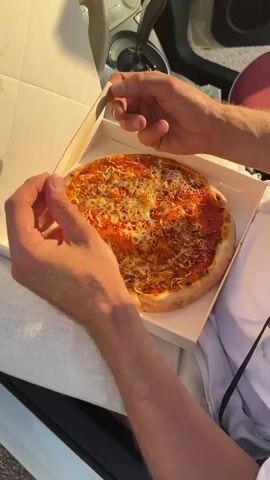 The video shows a person in a car holding a pizza with a small knife, about to cut it. The pizza appears to be a margherita, with tomato sauce and melted cheese. The person is wearing a white shirt, and the car's interior is visible in the background, including the dashboard and steering wheel. The scene transitions to an exterior shot of a "Gang of Pizza" vending machine, which dispenses pizzas 24/7. The machine is gray and black, with a screen displaying information and a payment slot. The vending machine is located in a parking lot, with trees and other vehicles visible in the distance. The overall tone of the video is casual and consumer-oriented, highlighting the convenience of the pizza vending machine.