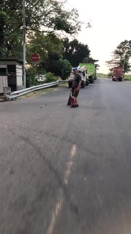 A man wearing a blue cap, grey t-shirt, and brown shorts stands on a paved road, holding a red dustpan and a broom. He appears to be sweeping debris from the road into the dustpan. Behind him, several large green trucks, likely garbage trucks, are parked in a line. The environment suggests an outdoor setting, possibly on the side of a road, with trees and vegetation visible in the background. The lighting indicates it may be either early morning or late evening.
