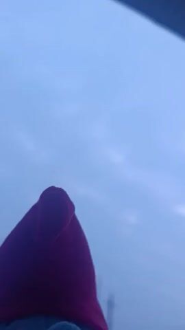 The video shows a close-up of a plush toy against a blue sky. The toy is light blue with a large nose, a smiling face, and a red pointed hat. The sky is a pale blue with some indistinct cloud formations. The camera appears to be angled upwards, capturing the toy against the sky.