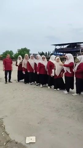 A group of women wearing maroon tops and hijabs stand in a line on a sandy beach. A man in a maroon t-shirt and black pants stands to the right of the group. In the background, there are several structures made of wood and what appears to be a large building with a dark roof. The sky is overcast.