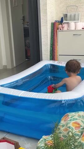 A toddler is playing in a blue and white inflatable kiddie pool outside on a tiled patio. The toddler is seated, shirtless, and is playing with a red and green toy truck. The child appears to be making car sounds. A partly open door is to the left of the pool, and a white storage unit with pink objects on top is visible to the right. A folded blanket is in the foreground and a small potted rosemary plant is next to it.