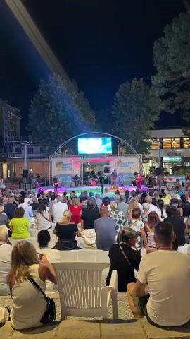 The video opens with a wide shot of an outdoor concert at night. A band is performing on a stage decorated with banners that say "Cattolica Mare Summer Village in Romagna." The band consists of a drummer, a keyboardist, and two guitarists, one of whom is also singing. A large LED screen behind the band displays colorful abstract imagery. In front of the stage, a tiered seating area is filled with an audience of people of various ages. The audience is seated and facing the stage, watching the performance. Trees frame the top of the scene, and lights illuminate the area. The video then zooms in slightly on the stage and the band, showcasing the performance. The camera remains focused on the stage and the audience's reaction for the duration of the clip.