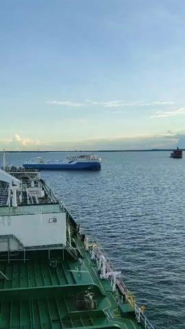 The video shows a view from the deck of a large ship, looking out at the ocean. Several other ships are visible in the distance. The sky is blue with some clouds, and the water is calm. The foreground of the video shows part of the deck of the ship, including green railings and metal structures. The middle ground features a blue and white ship. In the background, a red ship can be seen further out. The overall scene suggests a maritime setting, possibly during the daytime with good weather.