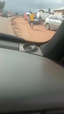 The video shows a view from inside a vehicle looking out at a dusty road with several vehicles and a person on a bicycle. The person on the bicycle is wearing a yellow shirt and is moving away from the camera. There is a silver SUV parked on the right side of the road. The road appears to be unpaved and there are other vehicles, including a white van and a red truck, further down the road. The sky is cloudy. A compass is visible on the dashboard of the vehicle in the foreground.