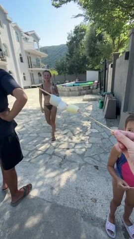 The video opens with a woman in a black bikini standing and holding a drink with marshmallows on a stick. A young girl in a pink and blue swimsuit stands to her right, holding marshmallows on a stick. A third person, partially visible, stands to the left. In the background, a house and trees are visible. As the video progresses, the focus shifts to the young girl and the woman behind her. They are roasting marshmallows over a grill. Another woman, with red hair and wearing a floral swimsuit, is also present. The scene appears to be outdoors on a paved area, possibly a patio or backyard, with a swimming pool in the background. The lighting suggests it is daytime.