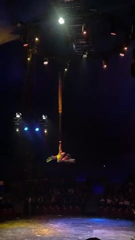 A circus performer is suspended upside down from a rope in the center of a dark circus tent. The performer is wearing a colorful costume and is performing an aerial stunt. The audience is seated in the background, watching the performance. The lighting is dim, with spotlights illuminating the performer and the circus ring. The overall atmosphere is one of excitement and awe.