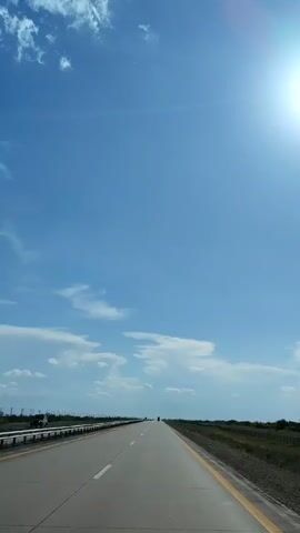 The video is a vertical, first-person perspective shot from a vehicle traveling on a highway on a clear, sunny day. The highway is paved and has yellow and white lines marking the lanes. A metal guardrail runs along the left side of the road, and a grassy shoulder with some vegetation borders the right side. In the distance, there are a few vehicles on the highway, and the horizon is flat with a sparse line of trees or structures. The sky is a vibrant blue with scattered white clouds. The sun is visible in the upper right corner, casting a bright glare. The overall impression is one of an open, expansive landscape and a long, straight road.