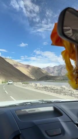 The video shows a view from inside a car driving on a road. The road is asphalt with white lane markings. A white car is visible ahead on the road. To the right of the road, there is a body of water, likely a river, with some vegetation along its banks. Beyond the river, there are large mountains with varied colors and textures, some illuminated by sunlight and others in shadow. The sky is blue with scattered white clouds. The dashboard and part of the windshield of the car are visible in the foreground. A yellow and orange decorative item is hanging from the rearview mirror. The camera is panning slightly to the right, following the road and the landscape.