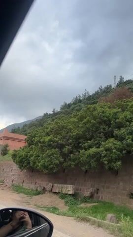 The video captures a view from inside a car driving along a dirt road in Taljarft, Morocco, under an overcast sky. In the foreground, a reflection of a person's hand holding a phone is visible in the car's side mirror. To the right, a long, rustic stone wall runs parallel to the road, featuring two doors: one light blue and another black with white geometric patterns. Bags containing materials are seen at the base of the wall. Behind the wall and ascending a hillside, several buildings with distinctive earthy-pink or terracotta hues are visible, blending with the natural landscape. Lush green trees and vegetation cover the hillside, and a Moroccan flag flies atop one of the buildings, confirming the location. The car is in motion throughout the recording.