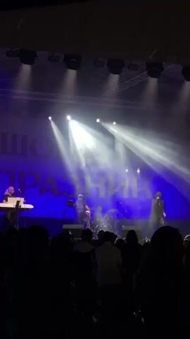 A video captures a live musical performance on a stage, viewed from the audience. Bright stage lights illuminate three male musicians against a large blue backdrop with white Cyrillic text. On the left, a man plays a keyboard. In the center, a man plays a bass guitar. On the right, a singer, dressed in black with a hat, performs into a microphone. A drum set is visible behind the bassist. The audience, silhouetted in the foreground, watches the performance.