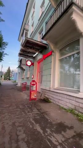 The video shows a person walking a dog on a leash down a sidewalk next to a building and a street. The building is light blue with a red sign that says "K&S". There is a red arrow pointing towards the entrance of the building. The dog is a light brown corgi. The street has cars and traffic lights. The sky is blue with some clouds. The weather appears to be pleasant.