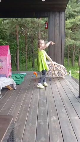 A young girl with blonde hair in a bright green dress and grey leggings dances on a wooden deck. She wears yellow Crocs and her hair is in a braid. To her left is a pink playhouse. To her right, an umbrella lies open on the deck. Behind her, a fire extinguisher is mounted on a wooden wall. The deck is surrounded by trees and vegetation. The girl is moving her arms and legs as if dancing, and she appears to be looking at someone off-camera. The video is filmed from a low angle.