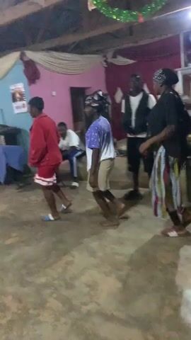 The video shows a group of four individuals and a toddler dancing and socializing indoors. The three individuals dancing in the foreground are wearing casual clothing and sandals, while the fourth person is seated in the background. The toddler, dressed in a dark jacket and pink shoes, also dances. The room is decorated with streamers and features a worn concrete floor, blue and pink walls, and mismatched chairs. A doorway is visible in the background, and a speaker stands to the right. The atmosphere appears lively and informal.