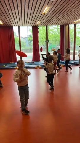 In a brightly lit indoor space, several young children practice spinning plates on sticks, under the supervision of an adult. The children, dressed in casual clothes, are scattered across the red floor, each attempting to keep their plate balanced. Some plates are red, others orange. The adult, wearing a black shirt and Adidas pants, moves among the children, offering guidance. Large windows with red curtains line one side of the room, offering a glimpse of trees outside. The ceiling is made of wooden slats. The atmosphere is active and cheerful, with the children focused on their juggling activity.