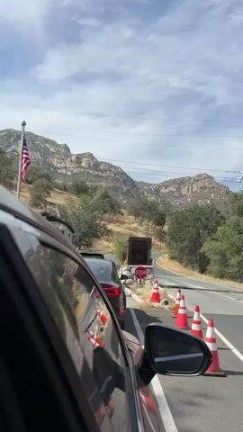 The video shows a line of cars stopped at a roadblock on a paved road. Orange and white traffic cones are set up along the side of the road, with a stop sign visible in the distance. An American flag waves from a flagpole on the left side of the frame. Mountains and trees are visible in the background under a cloudy sky. The camera is inside a vehicle, showing the side of the car and its side mirror. The cars are stopped, indicating a delay or closure.