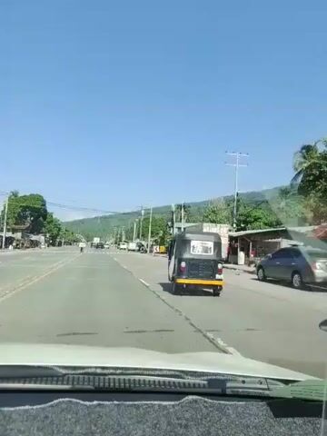 The video shows a view from inside a vehicle traveling on a road. The road is paved and has lane markings. On the right side of the road, there are several structures, including buildings and stalls. There are also utility poles with wires running overhead. A few vehicles are visible on the road, including a three-wheeled vehicle (likely a tricycle or auto-rickshaw), a white van, and a white truck. Trees and hills are visible in the background. The sky is clear and blue. The vehicle appears to be moving at a moderate speed. The video seems to be taken during daylight hours, suggesting a sunny day. There are no discernible interactions or activities involving people in the video. The overall impression is of a rural or semi-urban road scene in a tropical or subtropical region.