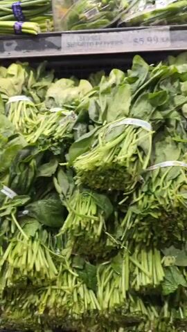 The video shows a close-up view of produce displayed in a grocery store. In the foreground, bundles of fresh green spinach are neatly arranged. Behind the spinach, several displays of root vegetables are visible, including turnips with purple tops and beets with reddish-purple skins. Further back, bags of peanuts in yellow mesh sacks are prominently featured. The lighting suggests an indoor retail environment, likely a produce section of a supermarket, with shelves and pricing labels partially visible. The camera moves smoothly, providing a clear view of the various food items.