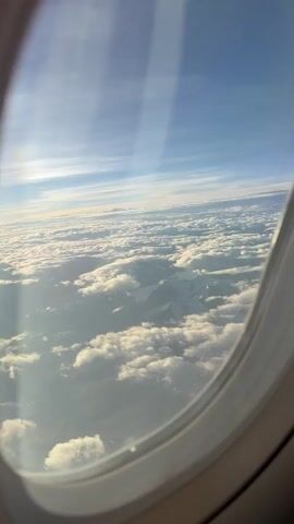 The video shows a view from an airplane window looking down at snow-capped mountains partially obscured by clouds. The sky above is blue with streaks of white clouds. The mountains appear rugged and mountainous, with white snow covering their peaks and slopes. The clouds are fluffy and white, forming a blanket over some parts of the mountains and the landscape below. The airplane window frame is visible in the lower right corner of the screen. The lighting suggests it is daytime, possibly early morning or late afternoon, due to the soft, angled sunlight.