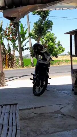 A black motorcycle with a black helmet on its handlebars is parked on a concrete surface. The motorcycle has a license plate that reads AE 5379 BC. Behind the motorcycle, there is a road and a field of green crops under a clear blue sky. Trees with green leaves are visible on both sides of the road. The scene is framed by a roof-like structure made of corrugated material and bamboo. The lighting suggests it is daytime.