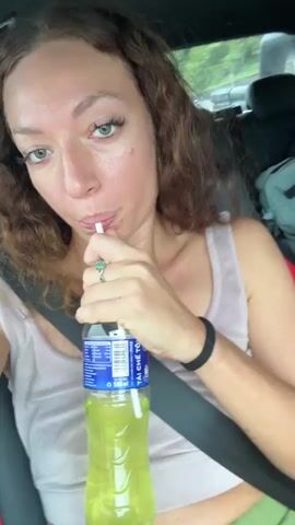 A woman with curly hair drinks from a bottle using a straw in the passenger seat of a moving car. She looks at the camera and purses her lips. The driver, a man wearing a pink shirt and a baseball cap, is visible. The car is driving on a rough, rocky road surrounded by greenery. The woman is wearing a seatbelt and a light-colored tank top. The bottle she is drinking from is clear with a blue label and contains a light green liquid.