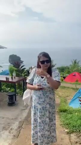 The video opens with a woman in a blue and white floral dress and sunglasses standing on a concrete patio, looking away from the camera and adjusting her hair. She holds a phone in her hand. Behind her is a view of the ocean with rock formations jutting out of the water. To the left, there's a dark wooden table with several small glass jars on it, topped with black stools. A large umbrella provides shade. To the right, there are several colorful tents pitched on a grassy area.

The camera then shifts to a wider shot, revealing another woman taking photos of the ocean view. The focus moves to the table and the ocean backdrop. The tents are visible on the periphery. The sky is overcast with white clouds. Palm trees and other greenery dot the landscape.