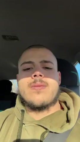 A man is in the driver's seat of a car, recording a selfie video. He has a buzz cut and a short beard, and is wearing a tan hoodie. The camera pans to reveal a Dalmatian-like dog sitting in the passenger seat, wearing a harness and secured with a seatbelt. The dog is looking out the window. The car appears to be in motion, with trees visible through the passenger window. There is music playing in the background.