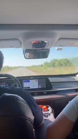 The video shows the interior of a car from the perspective of a rear passenger. The front passenger, with their back to the camera, is driving. Their right arm rests on the center console. The dashboard features two large digital screens displaying navigation and vehicle information. The windshield shows a two-lane road stretching into the distance, bordered by sparse vegetation and trees under a clear blue sky. The interior of the car appears to be modern, with leather seats and a panoramic sunroof. The overall impression is of a road trip on a sunny day.