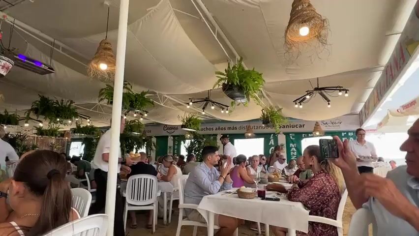 The video shows an outdoor restaurant with white tables and chairs under a white canopy. Several people are dining, and servers are bringing food. Hanging plants and woven light fixtures add to the ambiance. A server in a white shirt and black pants carries a tray of food. Another server, a woman in a white shirt, approaches a table with a tray. People are dressed casually, and the scene appears to be during the day. There is a sign in the background with text in Spanish.