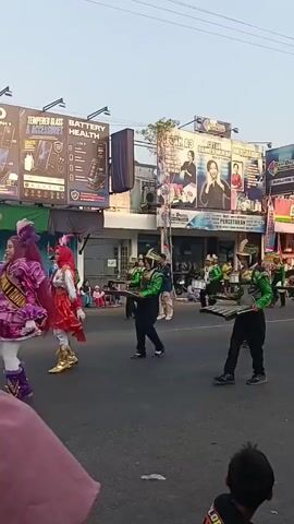 A marching band with participants wearing green and gold uniforms walks down a street. The participants are playing various instruments, including what appear to be xylophones and drums. There are spectators on the side of the road, including children and adults. Some spectators are sitting on the ground, while others are standing. The buildings lining the street have advertisements and signs. The sky is clear and the lighting suggests it is late afternoon or early evening.
