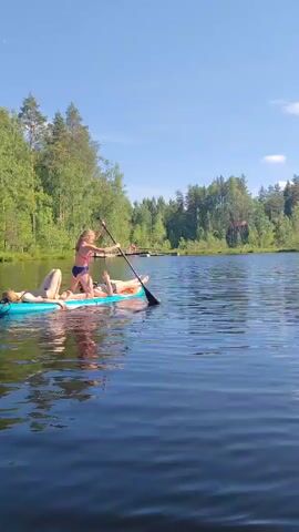 A bright, sunny day at a lake. A teal stand-up paddleboard is on the water, with two individuals on it. One individual, wearing a blue swimsuit and facing to the right, is kneeling on the board and paddling with a black paddle. Another individual, wearing a light-colored bikini, is lying on their back on the board, facing left. The board is surrounded by calm, dark blue water, and a lush, green forest with tall trees lines the distant shore. A wooden dock structure is visible in the water in the background. The sky is clear and blue.